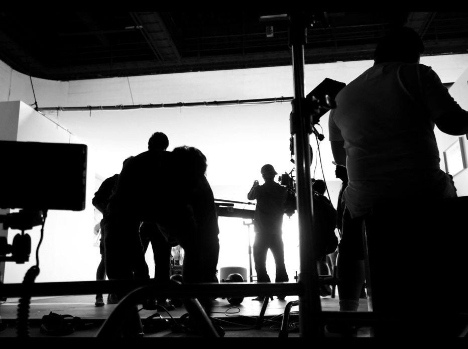 Behind the shooting video production and lighting set for filming which movie crew team working and silhouette shadow of camera and professional equipment in big studio for commercial advertising.